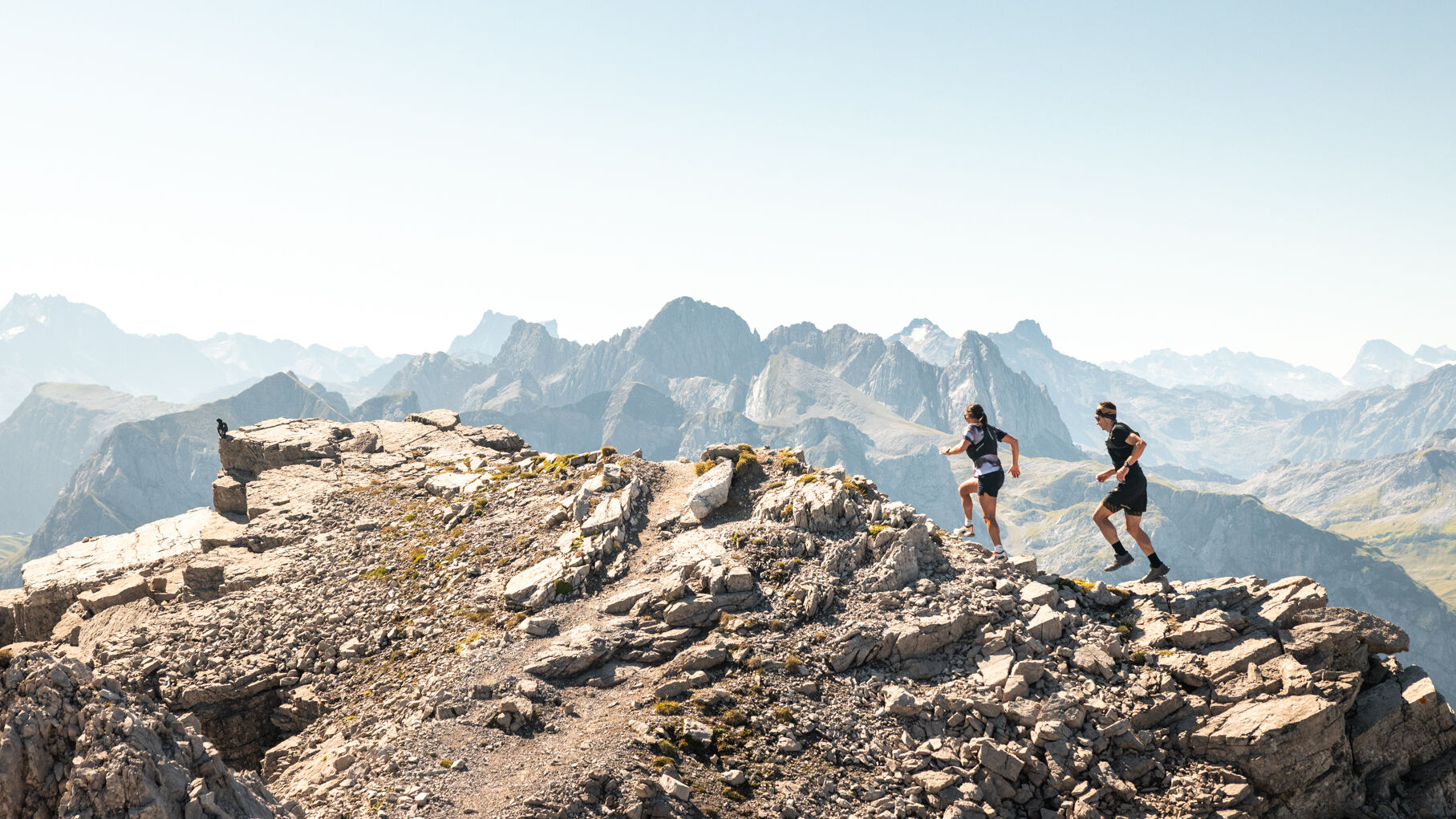 Zwei Trailrunner laufen durch eine steinige Landschaft und im Hintergrund sind Gipfel zu sehen.