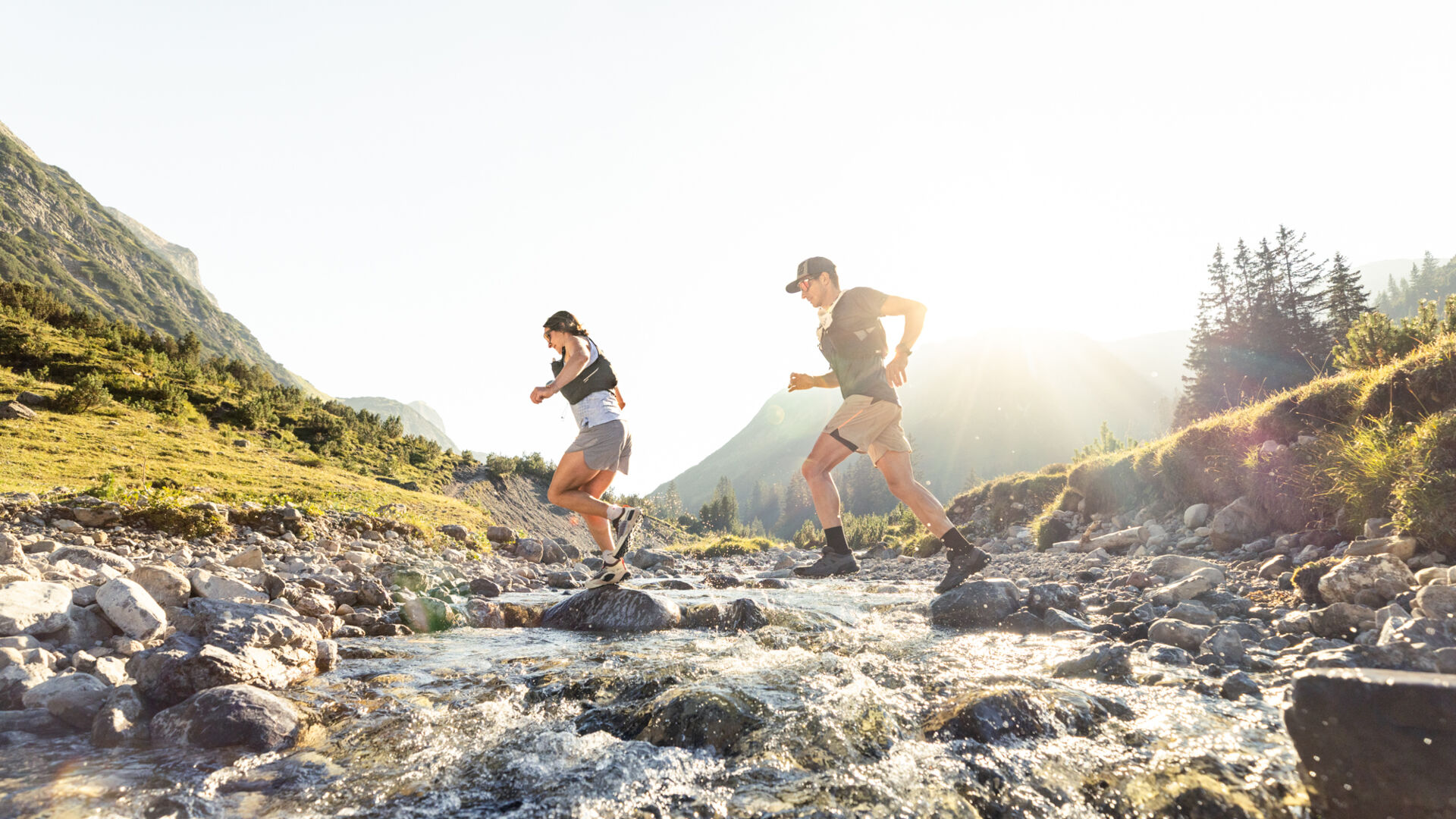 Zwei Trailrunner überqueren ein Gewässer und laufen durch die Berglandschaft