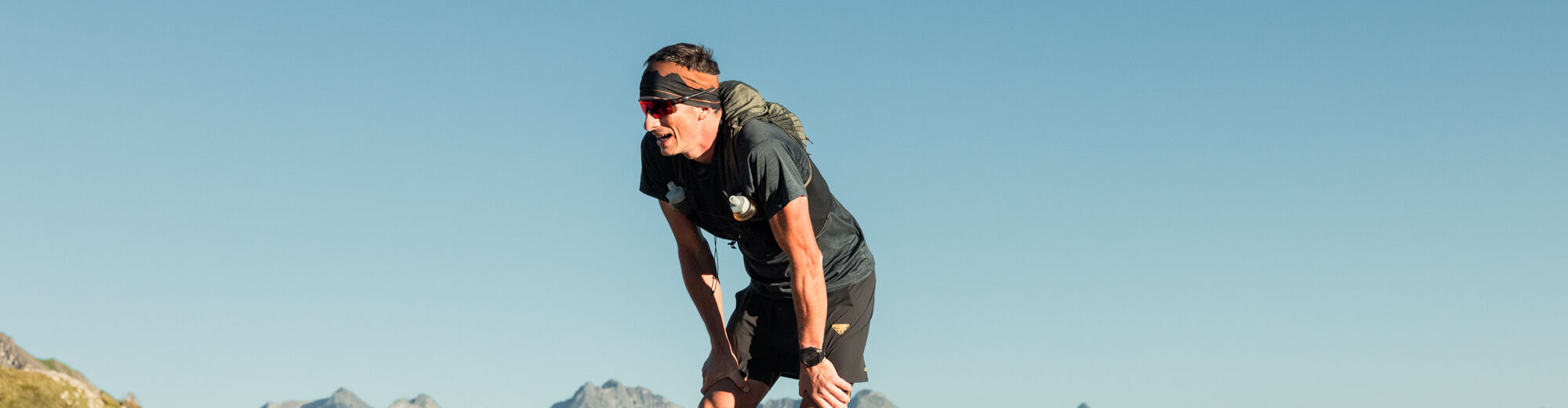 Erschöpfter Trailrunner mit Rucksack stützt sich auf einem Felsgipfel mit den Händen auf die Knie, im Hintergrund ein weites Panorama grüner Bergkuppen und schroffer Gipfelketten.