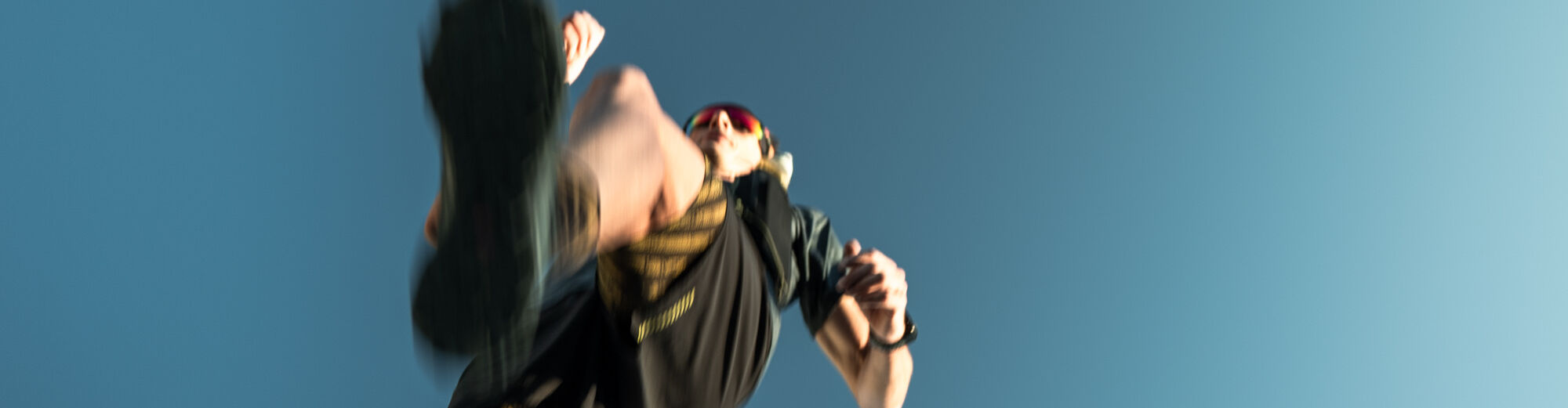 Dynamische Aufnahme aus der Froschperspektive: Trailrunner mit Sonnenbrille springt mit hoch angezogenem Bein durch die Luft vor blauem Himmel, starke Bewegungsunschärfe.