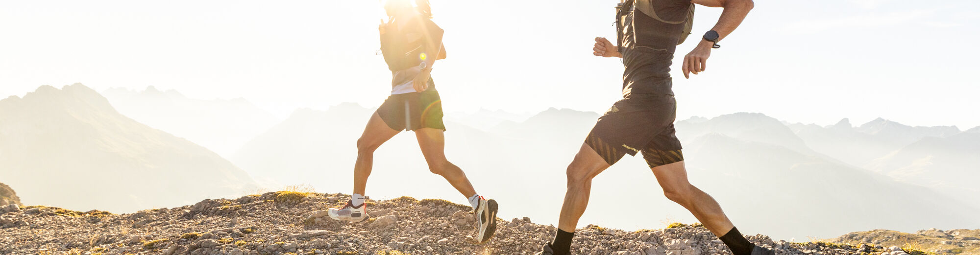 Zwei Trailrunner laufen vor einer Bergkulisse. Sie werden von Sonnenstrahlen geblendet