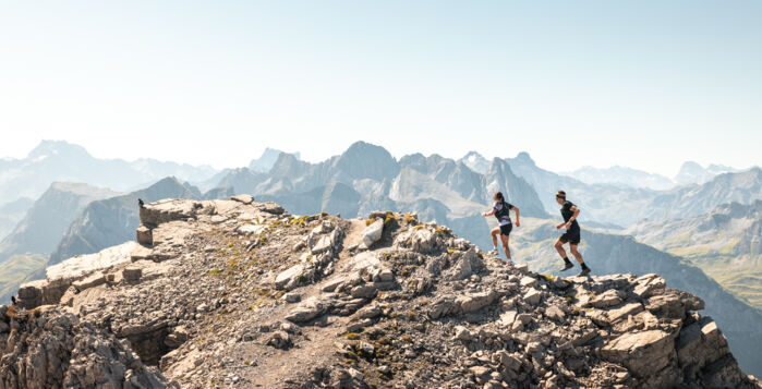 Zwei Trailrunner laufen durch eine steinige Landschaft und im Hintergrund sind Gipfel zu sehen.