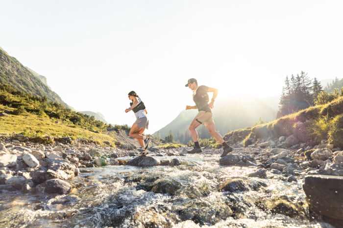 Zwei Trailrunner überqueren ein Gewässer und laufen durch die Berglandschaft