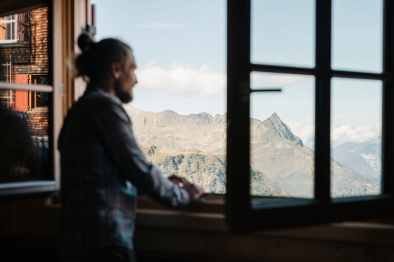 Wanderer blickt aus dem Hüttenfenster hinaus auf das umliegende Bergpanorama